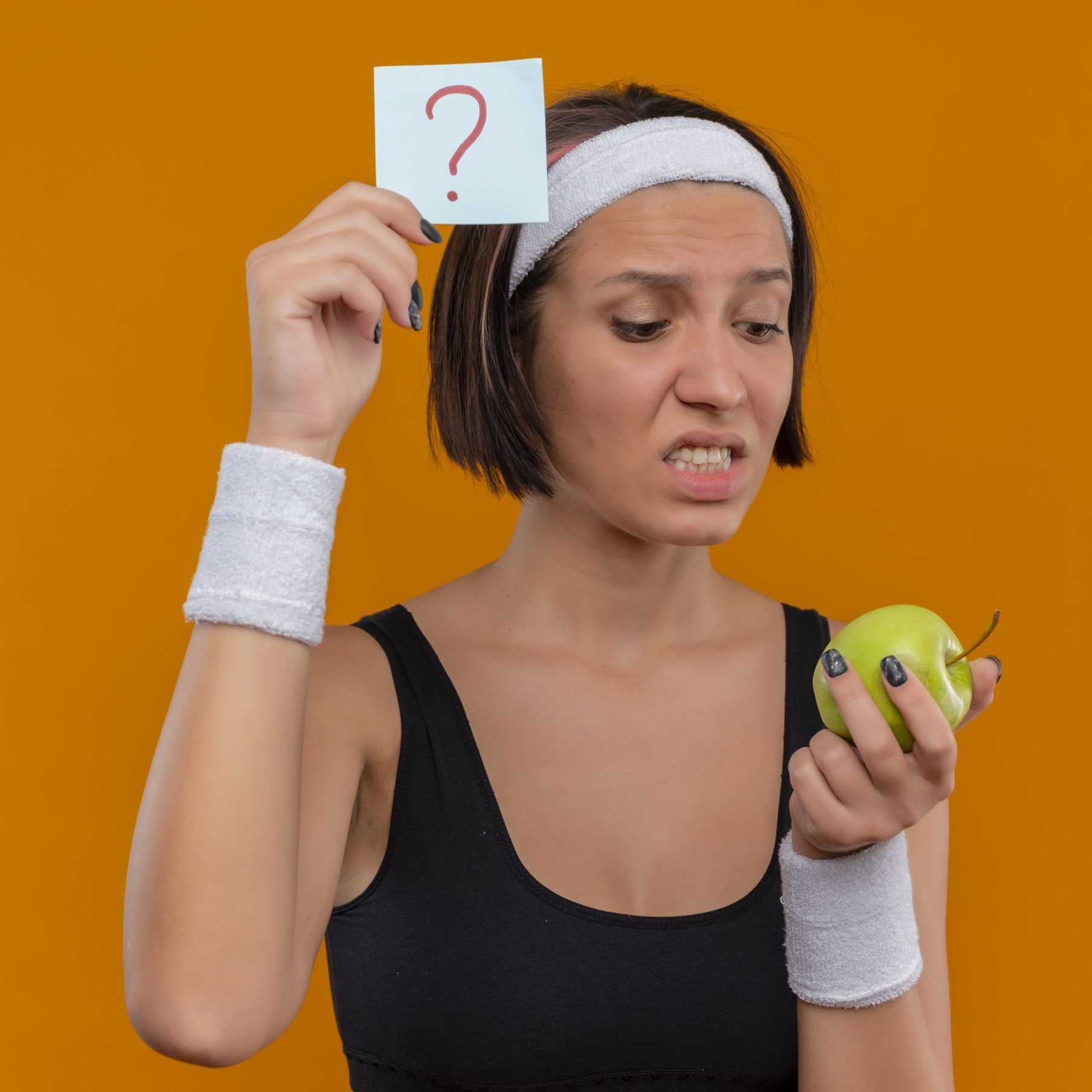young fitness girl in sportswear with headband showing reminder paper with question mark holding green apple looking at it confused and very anxious standing over orange background
